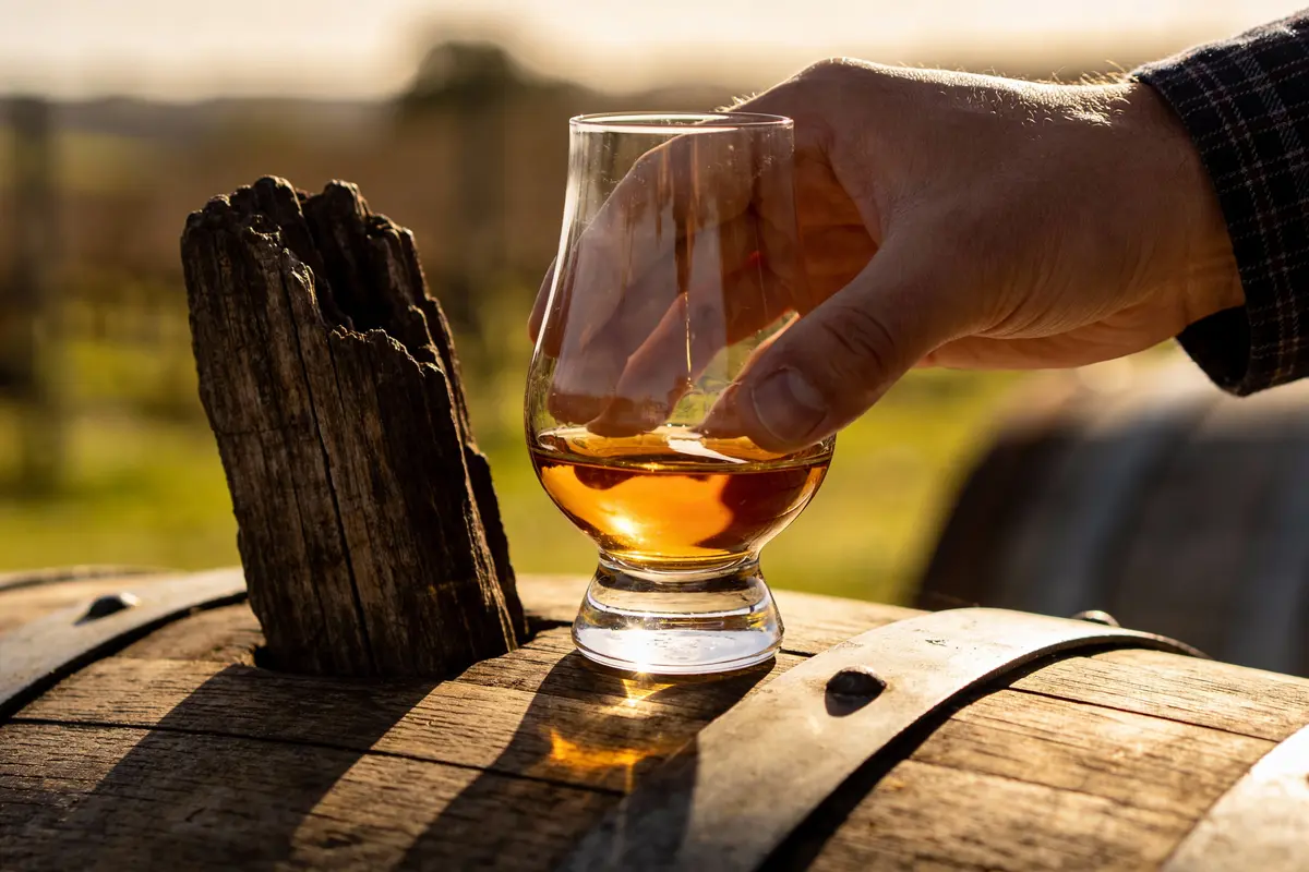 Main tenant un verre de whisky ambré sur un fût de chêne. Lumière dorée du soleil couchant sur la campagne.