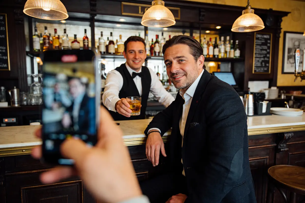 Client et barman souriants au bar, photographiés par un smartphone. Le barman tient une boisson à la main.