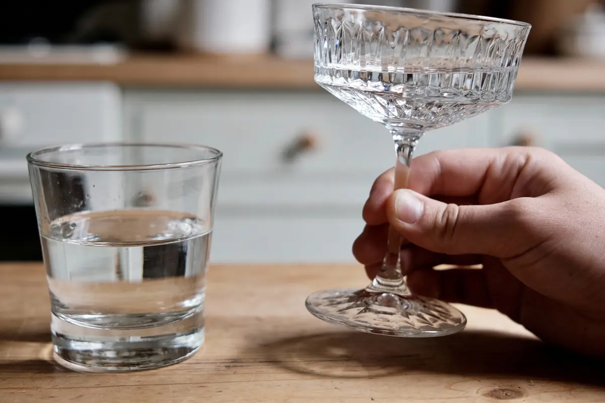 Une main tient une coupe d'eau élégante près d'un gobelet d'eau simple, tous deux sur une table en bois.