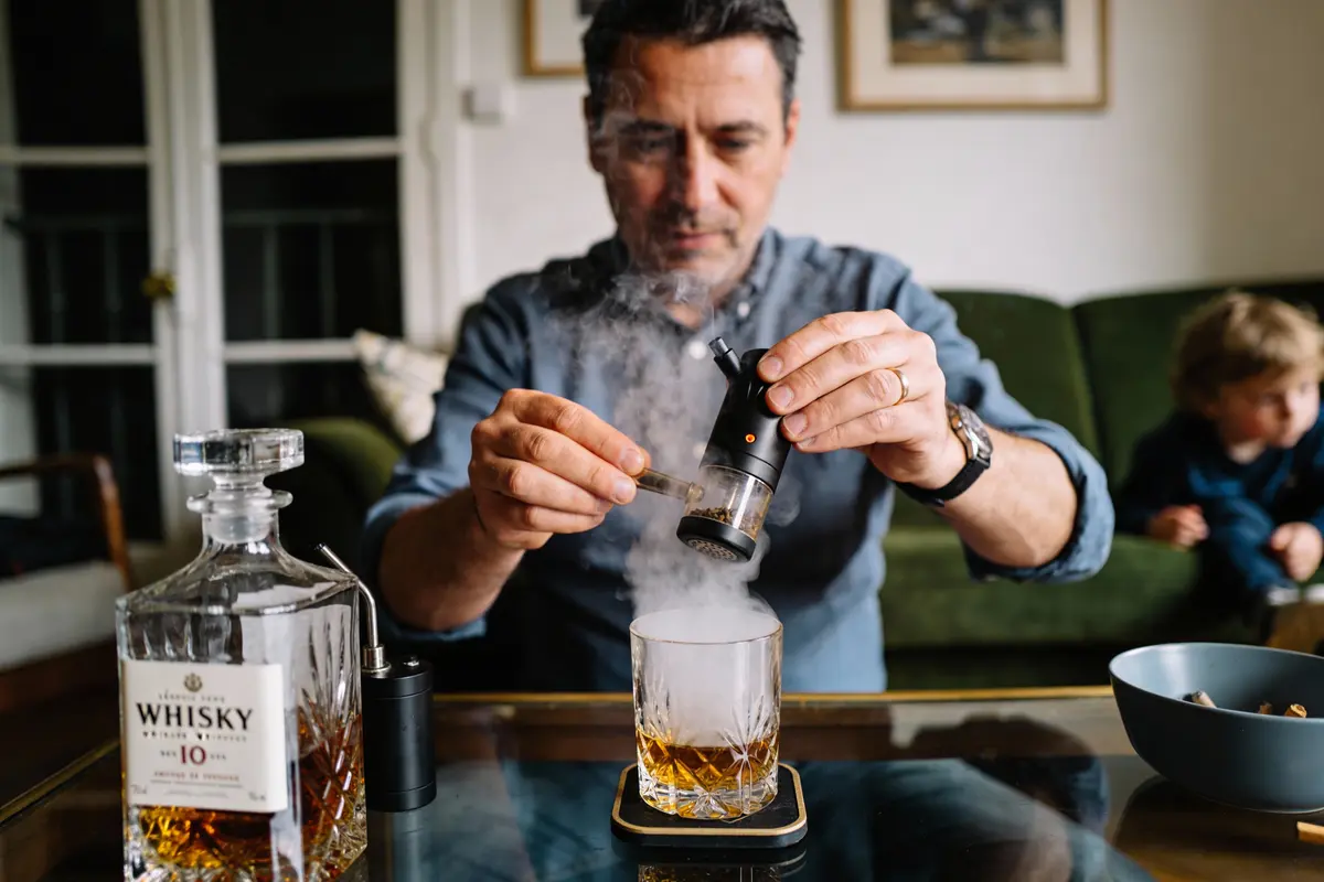 Un homme utilise un fumoir à cocktail pour infuser de la fumée dans un verre de whisky. Un enfant est en arrière-plan.