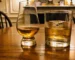 Deux verres de whisky sur une table en bois : l'un pour dégustation, l'autre avec glaçon. Ambiance intérieure chaleureuse.