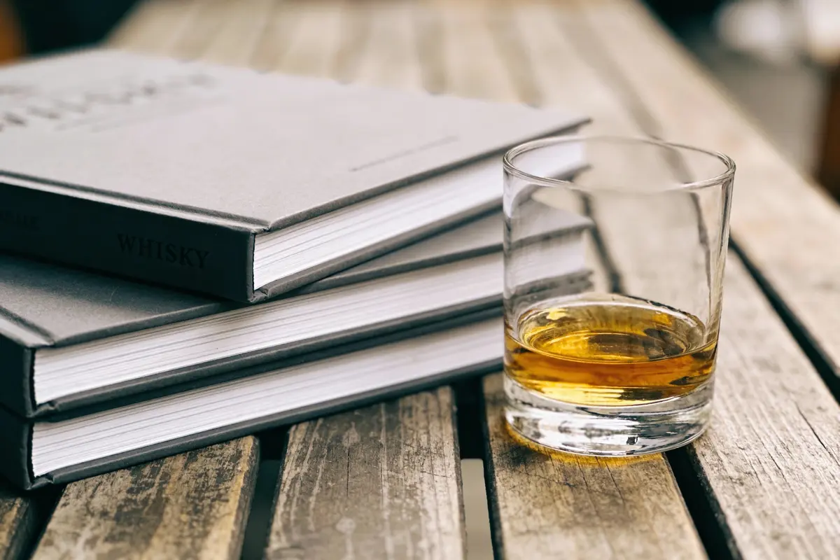 Une pile de livres gris, dont un intitulé "WHISKY", et un verre de whisky sur une table en bois rustique.