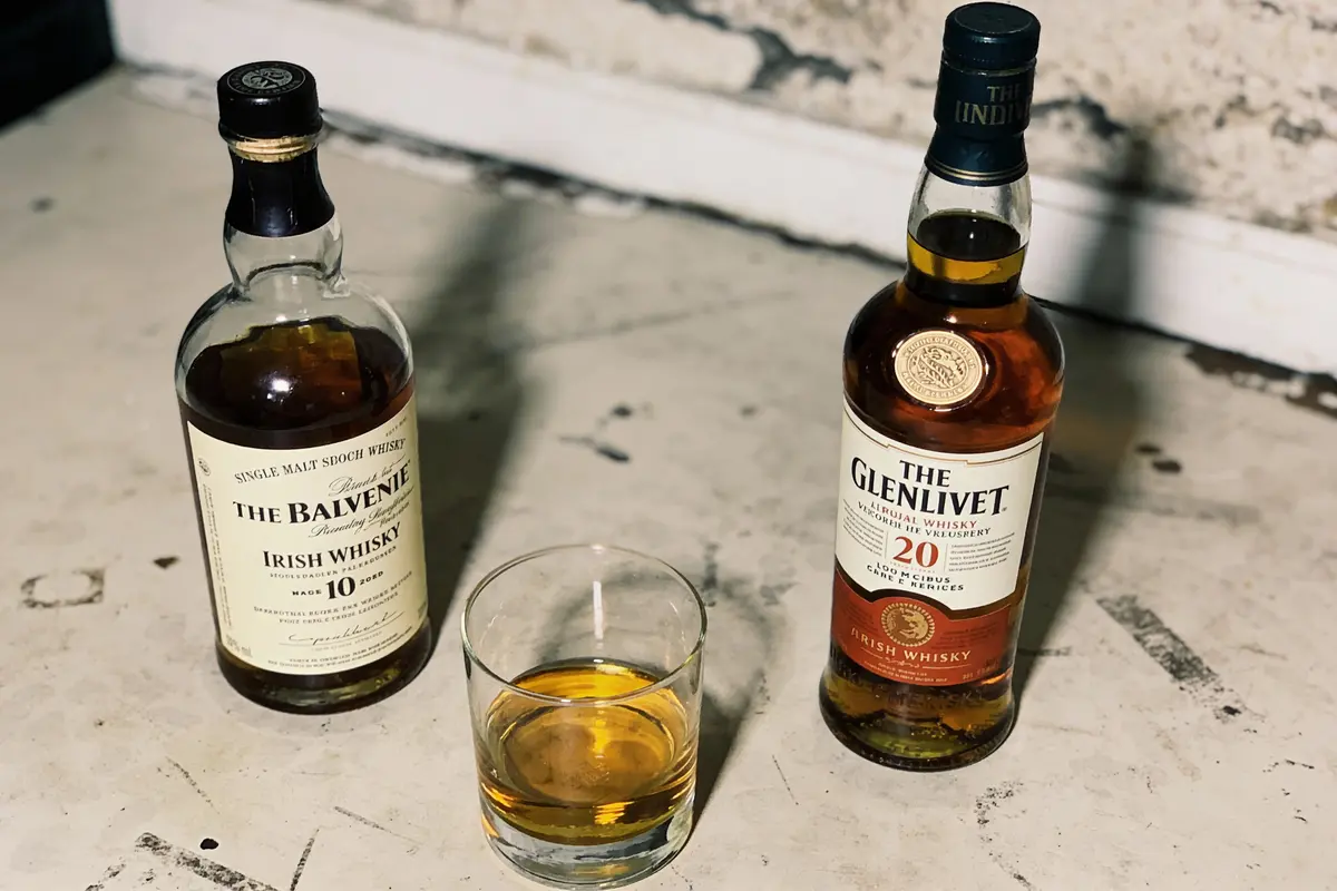 Deux bouteilles de whisky, Balvenie 10 ans et Glenlivet 20 ans, avec un verre de whisky sur une surface claire.