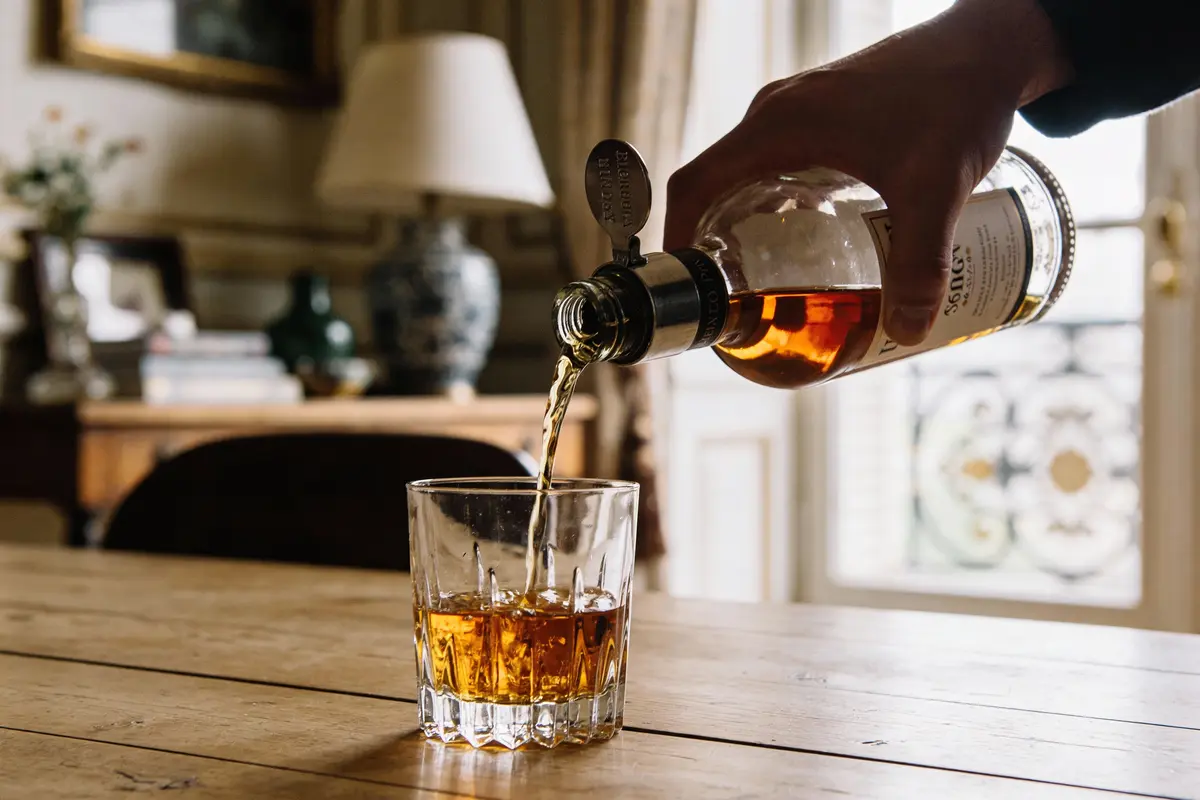 Une main verse du whisky d'une bouteille dans un verre sur une table en bois, dans un intérieur élégant flou.