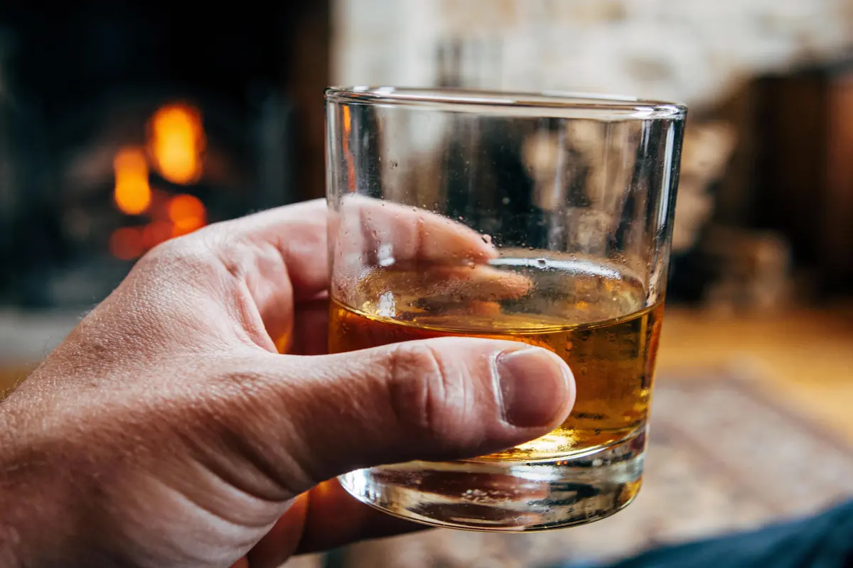 Voici une description précise et concise pour le texte alternatif : Main tenant un verre de whisky doré devant un feu de cheminée. Ambiance intérieure chaleureuse et relaxante.